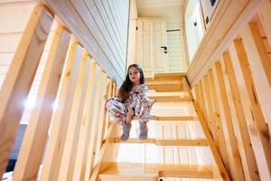 pequeño linda niña en pijama sentar en escalera en acogedor de madera minúsculo cabina casa. vida en campo. foto