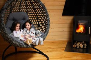 dos hermanas en pijama y de punto calcetines en cómodo colgando silla en contra hogar leyendo libros en acogedor de madera minúsculo cabina casa. vida en campo. foto