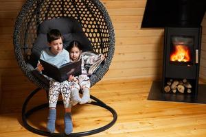 hermano con hermana leyendo libros en cómodo colgando silla en contra hogar en acogedor de madera minúsculo cabina casa. vida en campo. foto