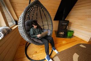 Teenager boy sit with laptop inside egg swing against fireplace at cozy indoors heating. photo