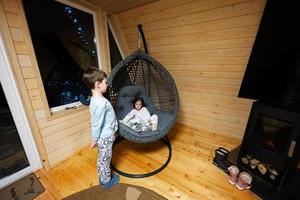 Brother with sister in comfortable hanging chair against fireplace in cozy wooden tiny cabin house. Life in countryside. photo