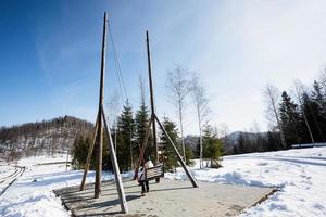 Back of mother swinging with daughters in big wooden swing in early spring snowy mountains. photo