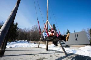 Mother swinging with daughters in big wooden swing in early spring snowy mountains against tiny house and car. photo