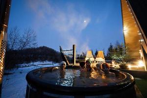 Family enjoying bathing in wooden barrel hot tub in the terrace of the cottage. Scandinavian bathtub with a fireplace to burn wood and heat water. photo