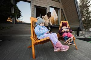 Two little girls sit on chairs at terrace off grid tiny house in the mountains and watching cartoons on mobile phones. photo