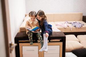 Two sisters are sitting on a couch, looking and read a book together. photo
