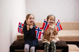 Two sisters are sitting on a couch at home with norwegian flags on hands. Norway children girls with flag . photo