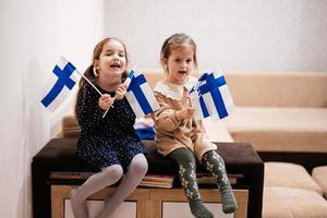 dos hermanas están sentadas en un sofá en casa con banderas finlandesas en las manos. Finlandia niños niñas con bandera. foto