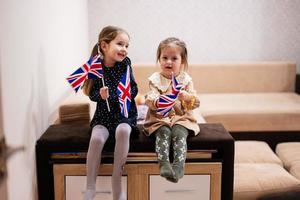 Two sisters are sitting on a couch at home with british flags on hands. Great Britain children girls with flag . photo