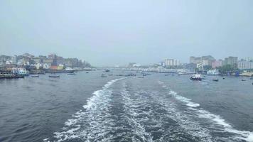 Wake of vessel. Wide wake trail from boat. Ships are standing both side of the river. River water.Cityscape from middle of the river. Small boats in the river. video