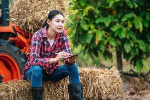 joven agricultora asiática sentada en un fardo de heno con una gran máquina tractora en tierras de cultivo. tecnologías modernas en gestión agrícola y concepto de agronegocios. foto