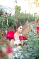 Young Asian woman wearing a white dress poses with a rose in rose garden photo