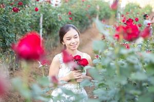 Young Asian woman wearing a white dress poses with a rose in rose garden photo