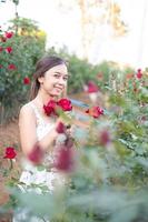 Young Asian woman wearing a white dress poses with a rose in rose garden photo
