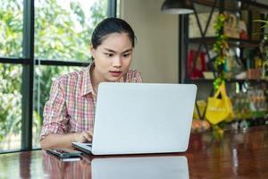 Cheerful young Asian woman using laptop while sitting on chair alone at coffee shop. Attractive female smiling with good news or chatting online in social network. Positive and Lifestyle concept photo