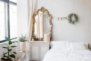 A double bed with white linens and pillows in the bedroom decorated for Christmas and New Year. Christmas wreath near the mirror at the dresser photo