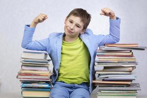 Happy smart school boy sits between stacks of books and shows a sign of strength. Overcoming learning difficulties. High school. photo