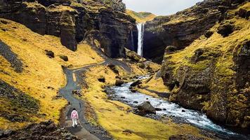 hembra turista caminar en ruta visitar famoso kvernufoos cascada punto de referencia. amarillo césped colinas en kvernufoss cascada. majestuoso verano ver de puro agua río en Islandia, Europa foto