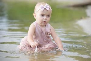 un hermosa pequeño niña se sienta en el río banco y obras de teatro con arena y agua. niño y naturaleza. foto