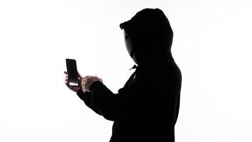 Hacker Anonymous and face mask with smartphone in hand. Man in black hood shirt holding and using mobile phone on white background. Represent cyber crime data hacking or stealing personal data concept photo