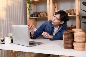 A young Asian man entrepreneur live streaming to sale craft products  to customers in his shop , Technology online business concept. photo