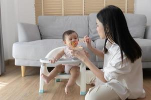 joven madre Ayudar bebé comiendo mezcla comida en bebé silla foto