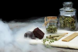 Pre-Roll cannabis joints and cake brownies with curing cannabis buds in a clear glass jar laying a black background photo