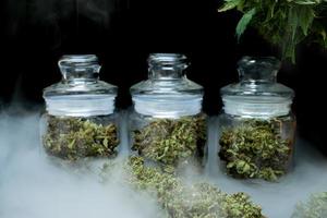 The marijuana buds in a clear glass jar sit on a smoky floor at the black background photo