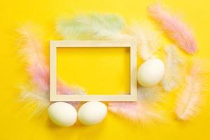 Easter white frame on a bright yellow background of chicken eggs and colored delicate feathers. Spring, religious holiday, Easter decoration, greeting, copy space, mock up photo