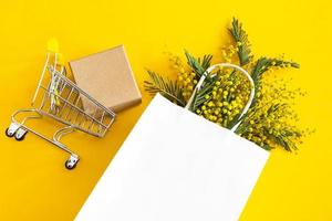 A bouquet of mimosa in a white mock up paper bag, craft gift box and a grocery cart. Spring shopping, gifts and promotions for International Women's Day. Yellow background, copyspace, flatlay.