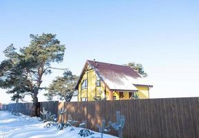 A yellow cozy house in the snow in winter in the village is surrounded by pine trees. Snow-covered roof, heating and ventilation pipes, trapezoid windows photo