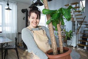 un contento mujer en un verde casa con un en conserva planta en su manos sonrisas, toma cuidado de un flor. el interior de un acogedor Respetuoso del medio ambiente casa, un hogar cocina, un pasatiempo para creciente y cría planta de casa foto