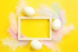 Easter white frame on a bright yellow background of chicken eggs and colored delicate feathers. Spring, religious holiday, Easter decoration, greeting, copy space, mock up photo