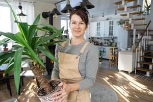 un contento mujer en un verde casa con un en conserva planta en su manos sonrisas, toma cuidado de un flor. el interior de un acogedor Respetuoso del medio ambiente casa, un hogar cocina, un pasatiempo para creciente y cría planta de casa foto