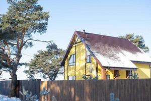 una acogedora casa amarilla en la nieve en invierno en el pueblo está rodeada de pinos. techo cubierto de nieve, tuberías de calefacción y ventilación, ventanas trapezoidales foto