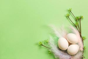 White Easter eggs in a delicate nest of colored feathers with unfurled leaf buds on the branches. Spring, a religious holiday, the birth of life. Copy space. Pale green background photo