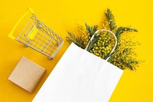 A bouquet of mimosa in a white mock up paper bag, craft gift box and a grocery cart. Spring shopping, gifts and promotions for International Women's Day. Yellow background, copyspace, flatlay.