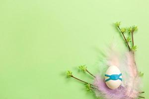 White Easter eggs in a delicate nest of colored feathers with unfurled leaf buds on the branches. An egg in blue plasticine medical mask is an epidemic, protection from the virus. Spring, Copy space. photo