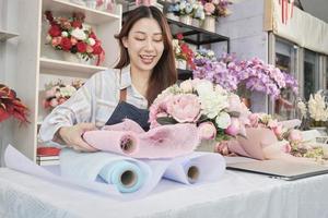 Young beautiful Asian female florist worker arranging bunch of blossoms on lovely valentine, happy work in colorful flower shop store with fresh bloom bouquets decorating, SME business entrepreneur.