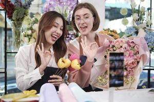 E-commerce business, two young female florist workers demonstrate and show floral arrangements via online live streaming with smartphone application in bright flower shop, a beautiful blossoms store.