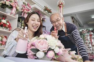 Old man and young beautiful Asian female florist workers water spraying a flora bunch together, happy work in a colorful flower shop store, fresh bloom bouquets decorating, SME business entrepreneur. photo