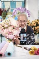 E-commerce business, one senior male florist workers demonstrate and show floral arrangements via online live streaming with smartphone application in a bright flower shop, a beautiful blossoms store. photo