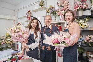 Portrait of flower shop workers team. Asian old male florist owner and young beautiful female employees in aprons with bunch of fresh floral bouquets arrangement for SME business, happy work in store.