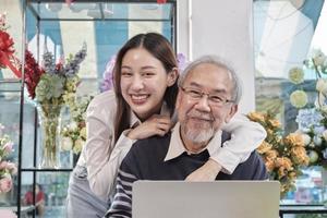 Asian young beautiful daughter teasing her father with love, senior male florist shop owner when working with laptop, happy work with smile and cheerful in colorful flower store, SME entrepreneur. photo