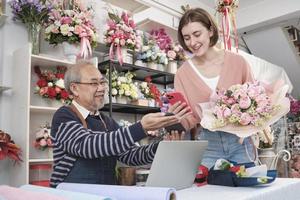 Cashless business entrepreneur. White customer woman shopping and digital payment by scanning a mobile phone application to an Asian elderly male florist owner. Beautiful floral shop, smart SME store. photo