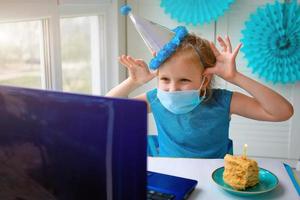 un contento pequeño chica, torcido en un médico máscara en frente de un ordenador portátil celebra un cumpleaños vía el Internet en cuarentena tiempo, autoaislamiento, en línea cumpleaños. foto
