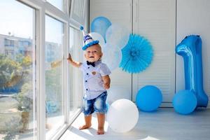 un pequeño chico en el gorra , soportes cerca un ventana panorámica, en su cumpleaños. 1 año viejo, globos foto