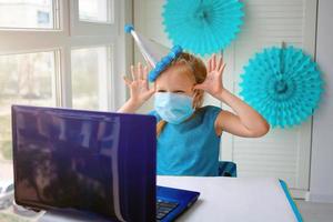 un contento pequeño chica, torcido en un médico máscara en frente de un ordenador portátil celebra un cumpleaños vía el Internet en cuarentena tiempo, autoaislamiento, en línea cumpleaños. foto