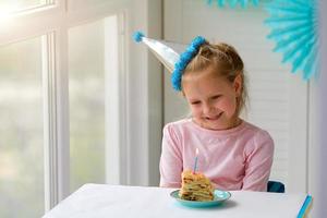 Happy little girl sits in a hat in front of a piece of cake and a candle. Birthday at home. photo