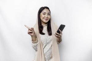 Excited Asian woman wearing hijab pointing at the copy space beside her while holding her phone, isolated by white background photo